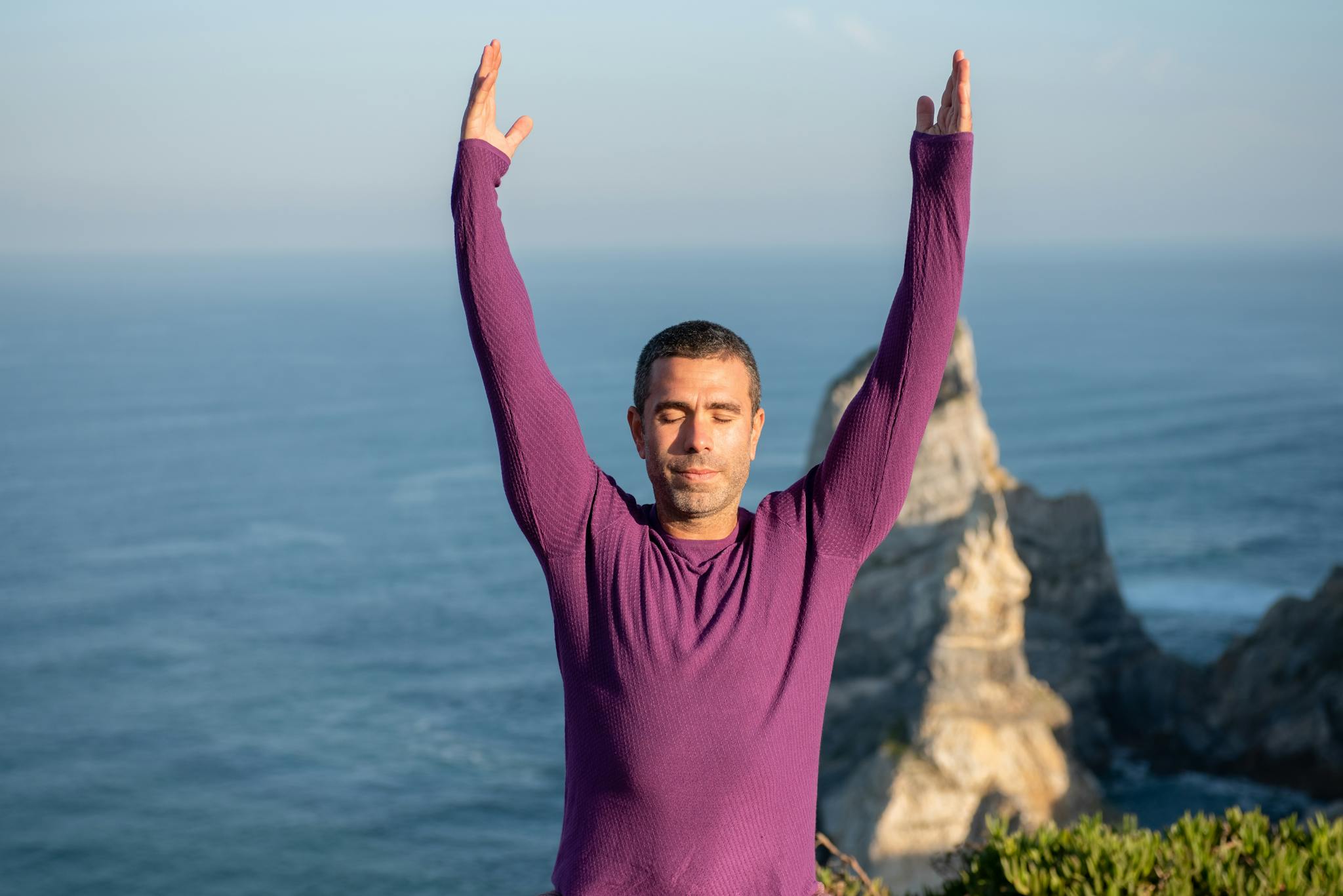 Man meditating with arms raised by the ocean cliffs in Portugal, wearing a purple top.